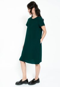 Rochie verde închis, până la genunchi, cu mâneci scurte și buzunare laterale, asortată cu încălțăminte neagră voluminoasă. Textură netedă și croi lejer.