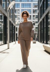 Beżowa, oversize bluza z okrągłym dekoltem i długimi rękawami, zestawiona z pasującymi luźnymi spodniami. Gładka faktura; widoczna biała metka.
