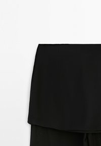 Jupe noire à couches avec une ceinture droite et un ourlet asymétrique, sur un fond clair uni.