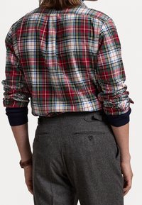 Camicia a quadri rossa, verde e bianca con bottoni, maniche lunghe e maniche interne blu, abbinata a pantaloni grigi con tasche posteriori.