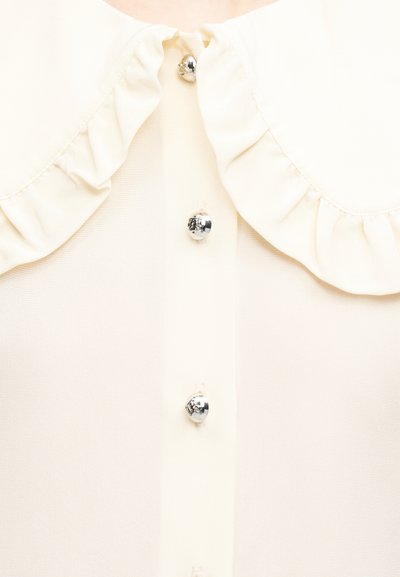 Κοντινό πλάνο κρεμ μπλούζας με βολάν στον γιακά και γυαλιστερά ασημί κουμπιά στο μπροστινό μέρος.