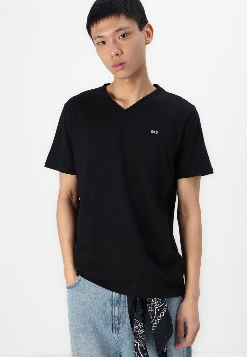 Schwarzes Baumwoll-T-Shirt mit V-Ausschnitt und kurzen Ärmeln; mit dezentem GAP-Logo auf der Brust, kombiniert mit hellblauen Jeans und einem schwarzen Bandana.