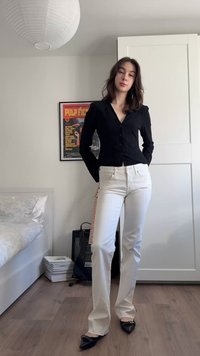 Γυναίκα με σκούρα μαλλιά, ντυμένη με μαύρο μπλουζάκι με κουμπιά, λευκό παντελόνι και μαύρα παπούτσια, στέκεται σε υπνοδωμάτιο με μια κιθάρα και αφίσα από το Pulp Fiction.