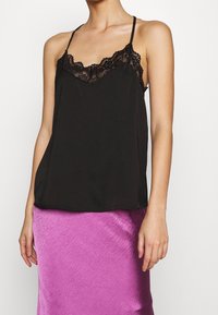 Femme portant un haut caraco noir orné de dentelle et une jupe en satin violet brillant, debout les bras détendus le long du corps.