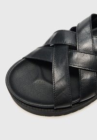 Sandal in pelle nera con design a strap intrecciato. Presenta una punta arrotondata e una suola interna testurizzata, montata su una suola flessibile nera.