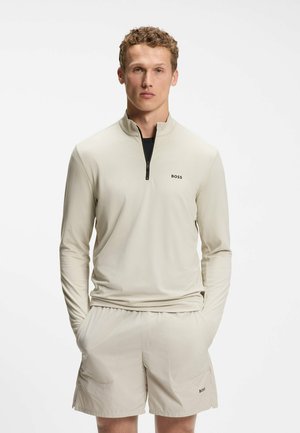 Giovane uomo con capelli ricci che indossa una maglia beige a maniche lunghe con cerniera al quarto e pantaloncini abbinati con loghi BOSS, in piedi con le mani in tasca.