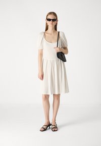 Robe blanche longueur genou avec des manches bouffantes, un corsage ajusté et une taille froncée. Portée avec des lunettes de soleil noires et un petit sac à main noir. Sandales noires.