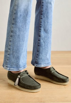 Persoon in blauwe jeans en donkergroene suède mocassin schoenen met crepezolen en kwastdetails op een houten vloer.