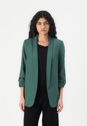 Green, non-structured blazer with a shawl collar, rolled sleeves, and an open front design, worn over a black top.