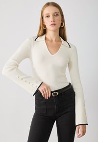 Ribbed white long-sleeve top with a V-neck, navy trim on the collar and cuffs, and gold button accents on sleeve hems, paired with black jeans.
