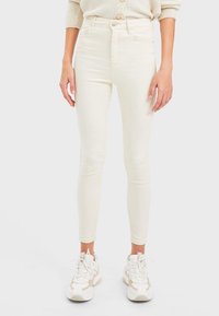 Jeans blancos ajustados de mezclilla elástica con cintura alta, que cuentan con un cierre delantero de botón y diseño de cinco bolsillos, combinados con zapatillas ligeras.
