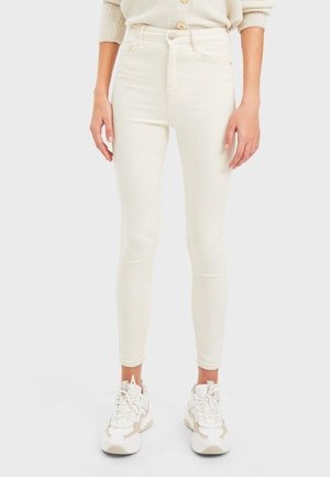 Jeans blancos ajustados de mezclilla elástica con cintura alta, que cuentan con un cierre delantero de botón y diseño de cinco bolsillos, combinados con zapatillas ligeras.