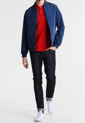 Blouson aviateur bleu marine porté sur un polo rouge, associé à un jean foncé et des baskets blanches. La veste possède une fermeture éclair et des poignets côtelés.