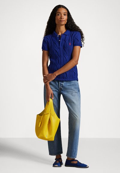 Modra polo majica z vzorcem pletenja, ovratnik, kombinirana z svetlo modrimi kavbojkami. Drži rumeno usnjeno torbico in nosi modre natikače.