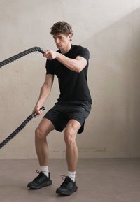 Camiseta atlética negra y pantalones cortos negros, zapatos de entrenamiento negros y calcetines blancos; individuo realizando ejercicio con cuerdas de batalla en interiores.