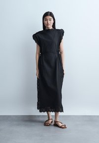 Γυναίκα που φοράει μαύρο midi φόρεμα χωρίς μανίκια με δαντέλα στα άκρα και καφέ σανδάλια με λουράκι στον αστράγαλο, στέκεται μπροστά σε έναν απλό ανοιχτό γκρι τοίχο.