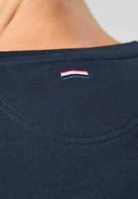 Navyblaue Baumwoll-T-Shirt-Kragen mit einem kleinen gestickten Trikolore-Logo (rot, weiß, schwarz) in der Nähe des hinteren Halsausschnitts. Glatte Textur und Nähte.