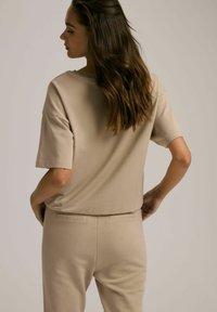 Beżowa bluzka z krótkim rękawem o luźnym kroju, wykonana z miękkiego materiału. Posiada okrągły dekolt i gładką strukturę. Widok z tyłu ukazuje prosty design.