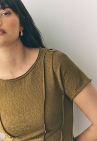 Olivgrön kortärmad t-shirt med rund halsringning och synliga sömmar, tillverkad av strukturerad tyg. Gyllene hoopörhänge synligt.