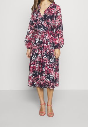 Vrouw draagt een midi wikkeljurk met lange mouwen met rode, witte en roze bloemenpatronen op een donkere achtergrond en koraalkleurige sandalen met enkelriempjes.