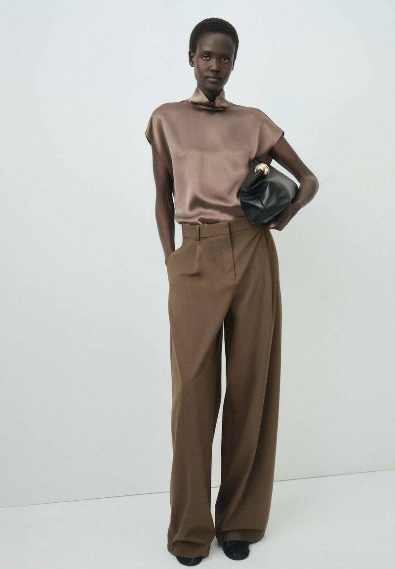 Bruine wijde broek gecombineerd met een zijden taupe top. Een zwarte handtas met een gouden accent wordt in één hand vastgehouden. Eenvoudige achtergrond.