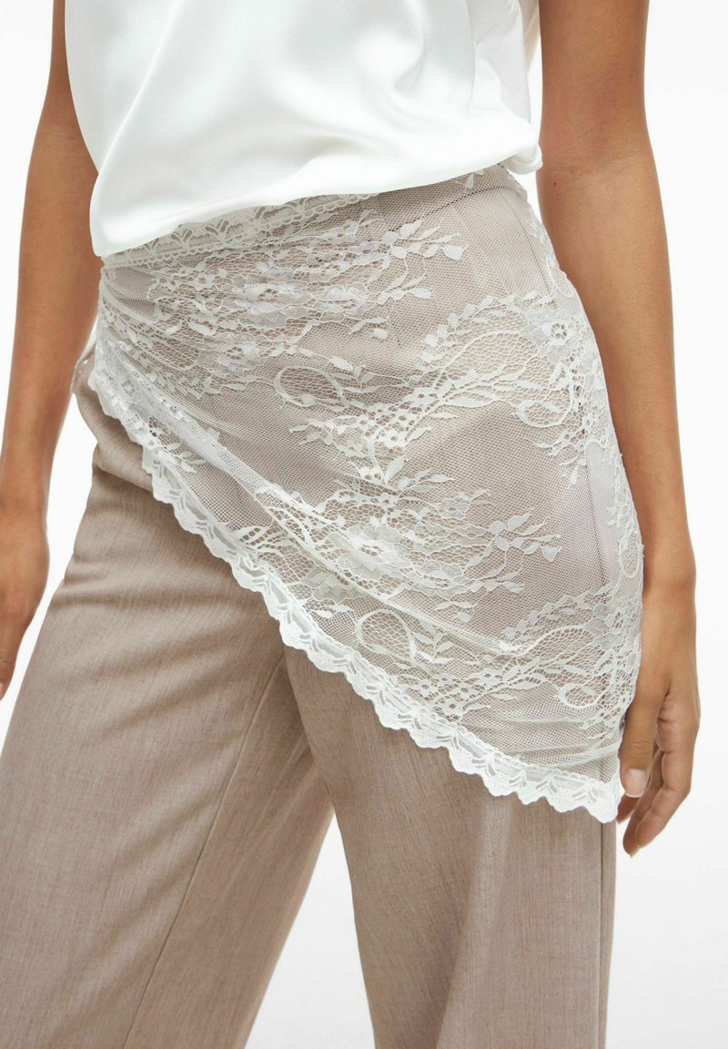 Lightweight tan pants paired with a sheer white lace overlay featuring floral patterns and scalloped edges. Smooth fabric texture.