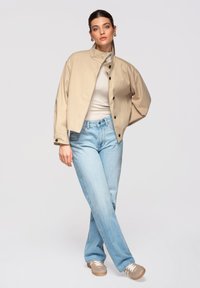 Femme portant une veste beige boutonnée, un haut blanc, un jean bleu clair et des baskets beiges, debout les jambes croisées devant un fond uni.