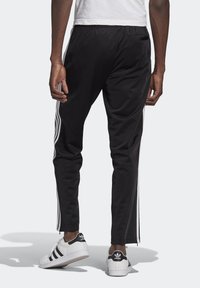 Pantaloni sportivi neri con strisce bianche sui lati, cintura elastica e vestibilità slim. Cerniera visibile alla caviglia. Indossati con sneakers bianche.