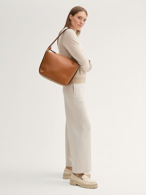 Donna che indossa un maglione e pantaloni beige, con una grande borsa a tracolla in pelle color marrone chiaro, in piedi contro uno sfondo chiaro e neutro.