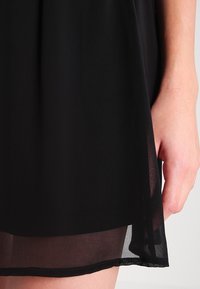 Händer vilar bredvid fållen av en svart kjol med ett genomskinligt underlager, som visar en del av ett ben mot en enkel ljus bakgrund.