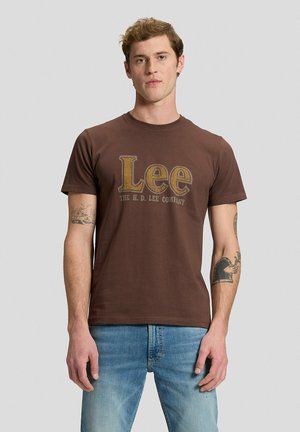 Uomo con capelli corti in piedi rivolto in avanti, indossa una t-shirt marrone con logo "Lee" e jeans blu, con tatuaggi su entrambi gli avambracci, sfondo neutro.
