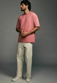 Helles rosa Baumwoll-T-Shirt mit entspannter Passform, Rundhalsausschnitt und kurzen Ärmeln, kombiniert mit cremefarbener gerade geschnittener Hose und weißen Sneakers.