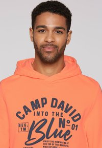 Sudadera naranja con un bolsillo tipo canguro en el frente. Presenta el texto "CAMP DAVID INTO THE BLUE" en azul oscuro. Hecha de una tela suave y texturizada.