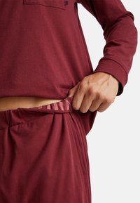 Osoba w burgundowej bluzce z długim rękawem i pasujących spodniach, podciągająca pas, aby pokazać paski wewnętrznego elastycznego detal.