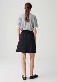 Frau mit braunen Haaren im Pferdeschwanz, die ein graues Kurzarmshirt, einen schwarzen knielangen Rock und schwarze Ballerinas trägt, steht vor einem einfarbigen Hintergrund.