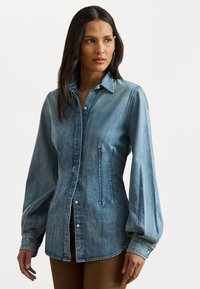 Ljusblå denimskjorta med långa puffärmar, knappad framtill och spetsig krage. Har sidfickor och en rundad nederkant.