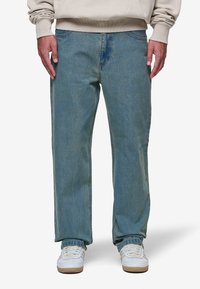 Jeans de lavagem azul claro, com um corte relaxado e acabamento desbotado, apresentando um design clássico de cinco bolsos e detalhes de costura, combinados com ténis brancos.