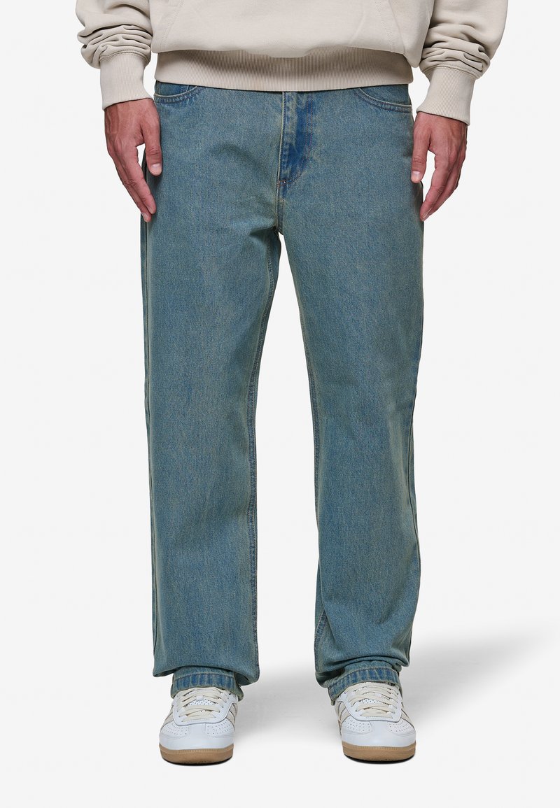 Jeans de lavagem azul claro, com um corte relaxado e acabamento desbotado, apresentando um design clássico de cinco bolsos e detalhes de costura, combinados com ténis brancos.