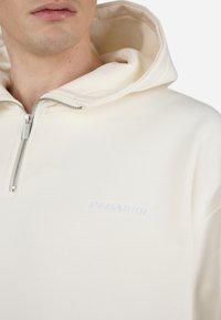 Cremefarvet zip-up hættetrøje med foret hætte, der har et hvidt broderet logo på brystet og en sølv lynlås. Blødt materiale, afslappet pasform.