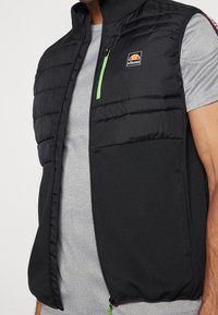 Czarna pikowana kamizelka z zielonymi akcentami na zamkach i naszywką z logo. Posiada gładką fakturę, wysoki kołnierz oraz dwie kieszenie z przodu.