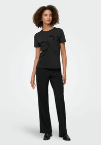 Ung kvinde med krøllet hår iført en sort kortærmet top med et struktureret sløjfedesign og sorte breddebenede bukser, stående.