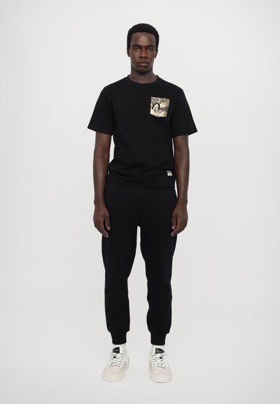 T-shirt en coton noir avec une poche à motif doré, associé à un pantalon de jogging noir et des baskets blanches ; design simple et décontracté.