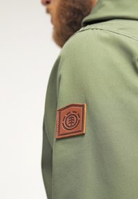 Veste en tissu vert avec un patch logo en cuir marron sur la manche gauche. Le design présente une coupe structurée et une texture subtile.