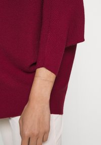 Gros plan sur le bras d'une personne portant un pull rouge à manches longues côtelé et un pantalon blanc, montrant la main et le poignet.