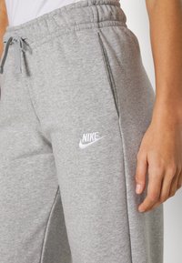 Gros plan sur la main d'une personne à côté d'un pantalon de survêtement gris Nike avec un cordon de serrage à la taille et un logo sur la cuisse gauche.
