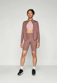 Lys brun treningstøysett som inkluderer en kortermet jakke, sportbh og høyt midje shorts. Matt tekstur, tettsittende design og logodetalj.