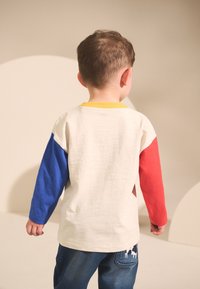 La maglietta a maniche lunghe presenta un corpo color crema, la manica sinistra blu e la manica destra rossa, con un colletto giallo. Texture morbida, design casual.