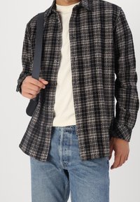 Homme portant une chemise à carreaux noire et blanche, un t-shirt blanc, un jean bleu, avec une bandoulière noire sur l'épaule.