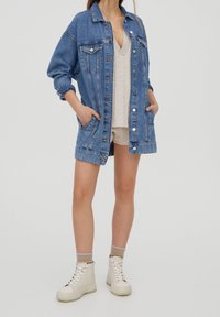 Veste en jean à lavage bleu, fermeture à boutons sur le devant, et deux poches poitrine. Portée sur un haut beige et un short, assortie à des baskets montantes blanches.
