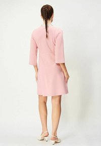 Rosa klänning med trekvartsärmar, slät tyg och en dekorativ krage. Bärs med vita, texturerade klacksandaler.